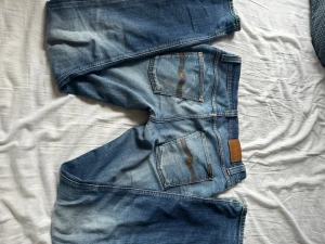 Blå jeans med kontrastsömmar - Säljer ett par klassiska blå jeans med snygga kontrastsömmar och femficksmodell. Jeansen har en tvättad look med slitningar och detaljerade bakfickor. Materialet är denim i bomull och passformen är rak. Perfekta för en avslappnad stil. Obs ett hål i bakfickan som syns på sista bilden.