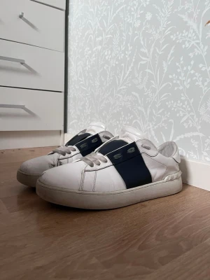 Valentino opens  - Säljer ett par vita valentino opens med bred marinblå rem över sidan. Skorna har klassisk låg profil, vita snören och tjock sula. Tillverkade i slätt läder och passar perfekt till grish eller casual outfits. Det är storlek 42 med passar 41 och 43 väldigt bra pris till denna sko..