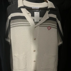 Casablanca tröja - Stickad skjorta med korta ärmar från Casablanca i vit och mörkgrå färg. Skjortan har knappar framtill, två fickor och ett broderat rött märke på bröstet. Snygg krage och coolt randigt mönster upptill ger en retro vibe.