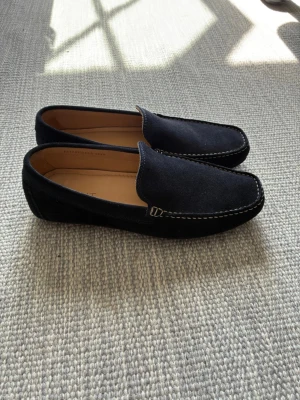 Mörkblå mockaloafers  - Snygga mörkblå loafers i mocka från Gant med ljus kontrastsöm runt tån. Skorna har en klassisk, stilren design och är fodrade med ljust läder invändigt. Perfekta för en clean och avslappnad look.