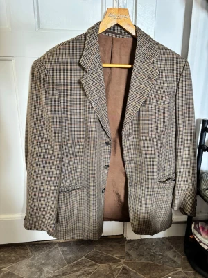 Brunrutig kavaj med klassiskt snitt - Snygg brunrutig kavaj med klassiskt snitt och två knappar framtill. Kavajen har två fickor med lock och en bröstficka. Fodrad insida i brunt tyg och diskret rutmönster i beige och mörkbrunt. Perfekt för en stilren look.