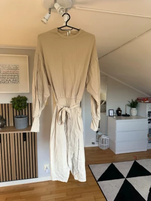 Beige långärmad klänning med knyt - Stilren beige klänning med långa ärmar och rund halsringning. Klänningen har ett knytband i midjan som ger en snygg siluett. Tillverkad i mjuk bomull, perfekt för en avslappnad men trendig look.