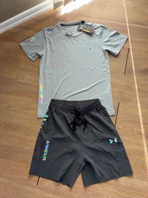 Under Armour träningsset (t-shirt + shorts) – helt nytt med tags - Säljer ett stilrent träningsset från Under Armour i grått – perfekt för gym, löpning eller kampsport.  ✔️ Helt nytt, oanvänt (tags kvar) ✔️ Lätt och snabbtorkande material ✔️ Bekväm passform ✔️ Stilren design  T-shirten har bra andningsförmåga och shortsen har elastisk midja med snörning för perfekt passform.  Säljes då det inte kom till användning.  Storlek: M/S Pris: 699 Skick: Nytt