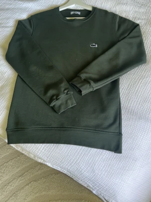 Mörkgrön sweatshirt från Lacoste - Mörkgrön sweatshirt från Lacoste med klassisk rund halsringning och broderad krokodillogga på bröstet. Tröjan har långa ärmar och ribbade muddar vid ärmslut och nederkant. Perfekt för en avslappnad och stilren look.