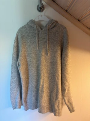 Ljusgrå Kashmir hoodie - En stilren ljusgrå hoodie med huva och dragsnören. Tröjan har ribbade muddar vid ärmslut och nederkant, och är tillverkad i ett mjukt, stickat material som känns riktigt skönt mot huden. Perfekt för en avslappnad och trendig look.