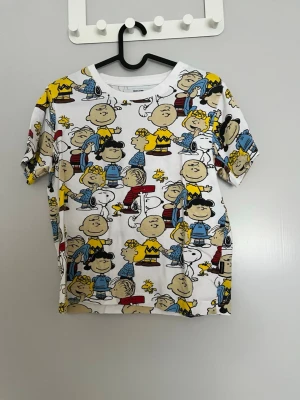 Peanuts t-shirt med färgglatt tryck - Rolig t-shirt med Peanuts-karaktärer i färgglatt all-over-tryck. Basen är vit och trycket har inslag av gult, blått, rött och svart. T-shirten har rund hals och korta ärmar, perfekt för dig som gillar serier och vill sticka ut.
