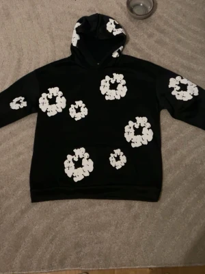 Svart hoodie med vita blommor - Säljer en svart hoodie med märket denim tears  Hoodien har en klassisk passform och är tillverkad i mjukt material som passar perfekt för en avslappnad stil. Perfekt för dig som vill sticka ut lite extra.