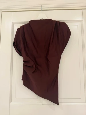Asymmetrisk vinröd blus - Snygg vinröd blus med asymmetrisk design och kort ärm. Blusen har en unik drapering och dragkedja i ryggen. Tillverkad i ett slätt tyg som ger en elegant känsla. Perfekt för dig som vill sticka ut med en cool och modern look. Har även paddade axlar, men gör inte att man ser stor ut!