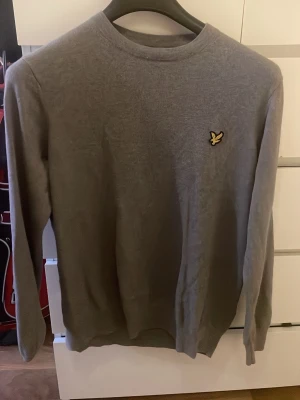 Grå stickad tröja från Lyle & Scott - Snygg grå stickad tröja från Lyle & Scott med rund hals och klassisk gul logga på bröstet. Perfekt basic-plagg med lång ärm och mjukt material som passar till det mesta. Enkel och stilren design.