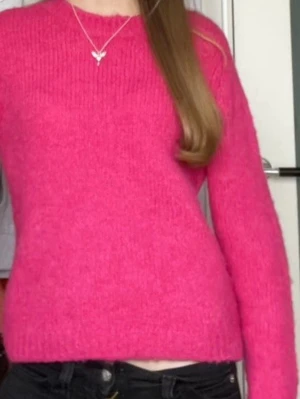 Stickad rosa tröja med rund hals - En stickad tröja i starkt rosa färg med rund halsringning och långa ärmar. Tröjan har en klassisk rak passform och ribbstickade muddar vid ärmslut och nederkant. Perfekt för att liva upp garderoben med en färgklick.