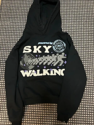 Svart hoodie med tryck 'Sky Walking' - Svart hoodie från Model A Club med stor grafisk print på bröstet där det står 'Sky Walking' och motiv av jordklot, stjärnor och silhuetter. Hoodien har huva och ribbade muddar. Perfekt för dig som gillar streetwear och unika prints.