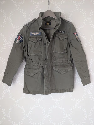 Polo Ralph Lauren Iconic Field Jacket  - Parkajacka vår/sommar från Polo Ralph Lauren med coola patchar på ärmar och bröst. Jackan har fyra stora fickor, dragsko i midjan och militärinspirerad design. Perfekt för dig som gillar utility och streetwear-vibe. Obs barn/ungdom.