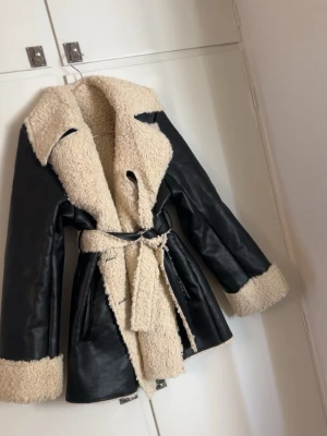Teddy kappa från NA-KD - Snygg svart kappa i faux läder med krage, ärmslut och insida i beige teddyfoder. Kappan har ett matchande bälte i midjan och stora slag. Perfekt för kalla dagar och ger en cool vintage vibe.