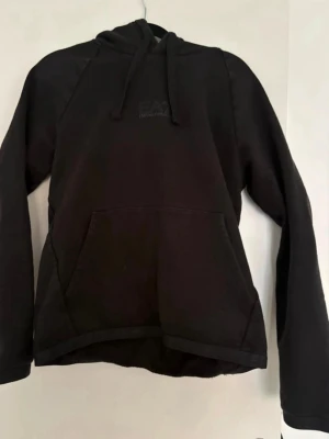 Svart hoodie från EA7 Emporio Armani - Säljer en svart hoodie från EA7 Emporio Armani med klassisk huva och dragsnören. Hoodien har en stor magficka och diskret logga på bröstet. Perfekt för en chill och stilren look. Materialet är mjukt och bekvämt, troligen bomullsblandning.