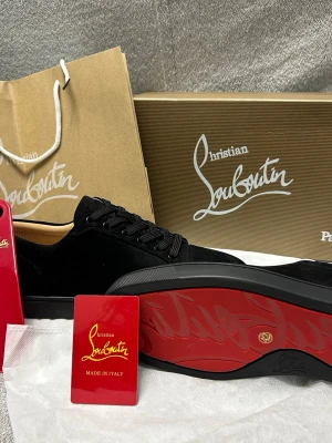 Christian Louboutin- Strl 42  - Stilrena svarta sneakers från Christian Louboutin i mocka med klassisk röd sula och svarta skosnören. Låg modell med diskret logga på innersulan och lyxig känsla. Perfekta för dig som vill ha något exklusivt och trendigt i garderoben. Kom privat för mer info de är helt nya och feta, oanvända och fraktas snabbt , vi kan även diskutera prisen samt prata mer om skorna 