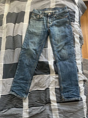 Blå jeans från Nudie Jeans ny pris på 1800kr (har kvitto - Säljer ett par klassiska blå jeans från Nudie Jeans med snygg slitning och raka ben. Jeansen har fem fickor, orange sömmar och Nudie-logga på bakfickan. Tillverkade i denim av bomull, perfekt för en avslappnad och trendig stil.