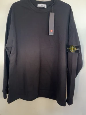  Stone Island - Säljer en helt ny  tröja från Stone Island med klassisk rund hals och ikonisk logotyp-patch på vänster ärm. Tröjan är i mjuk bomull och har en avslappnad passform. Perfekt för en stilren och trendig look.