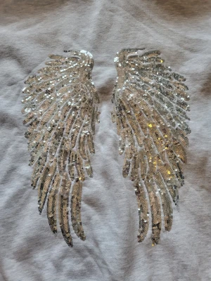 Grå hoodie med paljettvingar - Snygg grå hoodie med stora glittriga paljettvingar på ryggen. Hoodien har en klassisk passform med huva och ribbade muddar. Materialet ser ut att vara mjuk bomullsblandning, perfekt för en chill och trendig look.