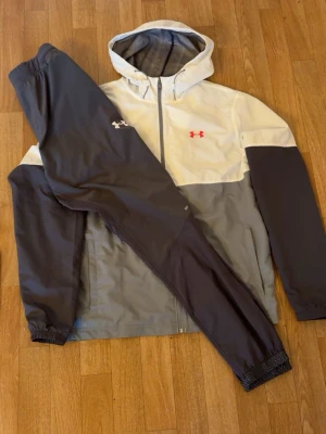 Under Armour träningsset grå/vit - Snyggt träningsset från Under Armour med jacka och byxor. Jackan är tvåfärgad i grått och vitt med huva och dragkedja, samt röda och vita loggor på bröstet. Byxorna är mörkgrå med resår i midja och benslut. Perfekt för träning eller sport.