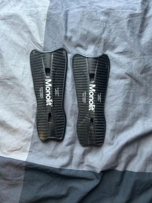 Monolit benskydd i kolfiber - Säljer ett par svarta benskydd från Monolit i kolfiber med snyggt mönster och vit logga. De är lätta, tunna och har en ergonomisk form som passar perfekt för fotboll eller andra sporter där skydd behövs.