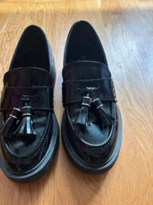 Svarta lackloafers med tofsar - Säljer ett par snygga svarta loafers i lackmaterial med dekorativa tofsar framtill. Skorna har en chunky sula och klassisk form som ger en stilren och trendig look. Perfekta för dig som vill ha en modern twist på klassiska loafers.