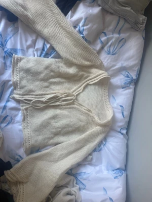 Krämvit stickad kofta med knytband - Söt krämvit stickad kofta med långa ärmar och knytband framtill. Koftan har en lätt croppad passform och är gjord i mjukt garn. Perfekt att slänga över en topp för en chill look.