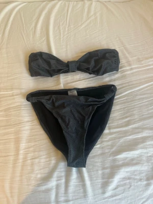 Mörkgrå bandeau bikini med små prickar - Snygg mörkgrå bandeau bikini med små prickar. Överdelen är axelbandslös med knäppning bak och en dekorativ knut framtill. Underdelen har en klassisk passform. Perfekt för stranden eller poolen nu till sommaren! Underdelen är i storlek S och överdelen i storlek M, men båda sitter som S.
