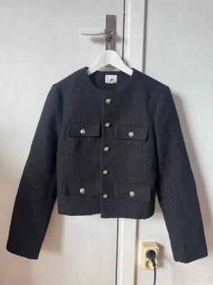Svart tweedkavaj med knappar - Stilren svart kavaj i tweed med fyra fickor framtill och dekorativa knappar. Rak, kortare modell med rund halsringning och långa ärmar. Perfekt för en trendig och klassisk look.