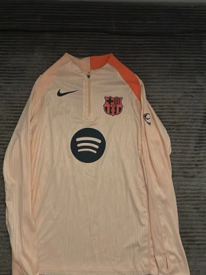 Barcelona träningströja Nike beige - Snygg beige långärmad träningströja från Nike med FC Barcelona-logga och Spotify-sponsor på bröstet. Tröjan har dragkedja vid halsen och orange detalj på axeln. Perfekt för fotbollsträning eller sportiga aktiviteter.