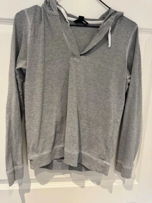 Grå hoodie från H&M med huva - Säljer en stilren grå hoodie från H&M med huva och vita dragsnören. Hoodien har långa ärmar, mjukt material och en avslappnad passform. Perfekt för en chill look och enkel att matcha med jeans eller joggers.