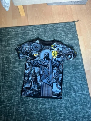 Svart Brasilien t-shirt Nike - Cool svart t-shirt från Nike med Brasilien-tema och detaljerad grafik av Kristusstatyn och andra motiv. T-shirten har korta ärmar, rund hals och gula loggor. Perfekt för dig som gillar unika prints och sportig stil.