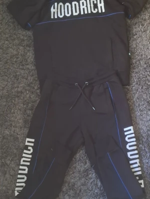 Svart set från Hoodrich med blå detaljer - Snyggt svart set från Hoodrich med matchande hoodie och byxor. Setet har vita tryck med Hoodrich-logga på både tröja och byxor samt tunna blå linjer som detaljer. Hoodien har huva och magficka, byxorna har dragsko i midjan. Perfekt för en streetwear-look. 1200 som orginalpris.