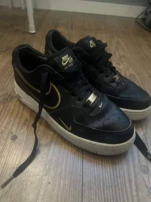 Nike Air Force 1 low ’07  - Snygga Nike Air Force 1 sneakers i svart läder med gulddetaljer på logga, snörning och tungan. Klassisk vit sula och lågt skaft. Perfekta för dig som vill ha en clean och stilren look med lite extra bling.