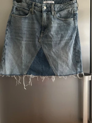 Jeanskjol från Calvin Klein Jeans - Trendig jeanskjol från Calvin Klein Jeans i ljusblå denim med råa kanter och patchwork-detaljer i olika blå nyanser. Kjolen har klassiska fickor fram och bak samt bälteshällor. Perfekt för dig som gillar en edgy och avslappnad stil.St 28