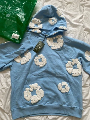Blå hoodie med vita moln från Denim Tears - Säljer en ljusblå hoodie från Denim Tears med unika vita molnliknande applikationer över hela tröjan. Hoodien har huva, magficka och ribbade muddar. Perfekt för dig som vill sticka ut med en kreativ och trendig look.