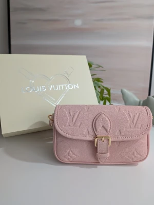 Iouis Vuitton Nano Diane🌸 - Denna nätta axelremsväska är utförd i en mjuk, puderrosa nyans. Materialet består av ett strukturerat läder med ett genomgående präglat mönster av ikoniska monogram och blommotiv, vilket ger ytan en tredimensionell känsla.Väskan visas tillsammans med en tillhörande förvaringsbox i en ljus benvit ton. Boxen pryds av ett stiliserat silverfärgat tryck föreställande en väska formad som en symbol för tillgivenhet, tillsammans med varumärkets namn i stora bokstäver.