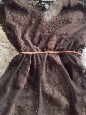 Brun babydoll spets top  från H&M - Snygg brun babydoll tröja från H&M i spets med blommigt mönster och transparenta partier. Klänningen har ett tunt bälte i midjan som markerar siluetten. Perfekt för dig som gillar romantiska detaljer och vill sticka ut.