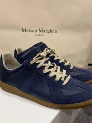 Maison Margiela blå sneakers - Snygga blå sneakers från Maison Margiela med beige snörning och klassisk gummisula. Skorna har en mix av mocka och läderdetaljer, låg profil och stilren design. Perfekta för dig som gillar minimalistisk och trendig streetstyle.