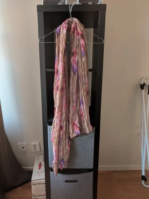 Beige sjal med blommigt mönster - Luftig och skrynklig beige sjal med blommigt mönster i rosa och lila nyanser. Perfekt att slänga över axlarna för en färgklick till outfiten. Tunt och mjukt material som känns lätt mot huden.