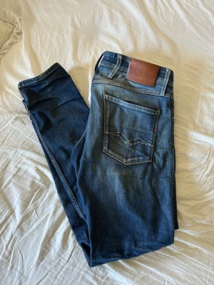 Exklusiva Replay Anbass - Säljer ett par blå jeans från Replay med slim fit (anbass) och klassisk femficksdesign. Jeansen har tagits väldigt väl hand om och har sköts ordentligt. Jeansen har snygga slitningar och en brun läderpatch bak i midjan. Materialet är stretchigt denim som sitter  bekvämt. Perfekta för en trendig och avslappnad look.