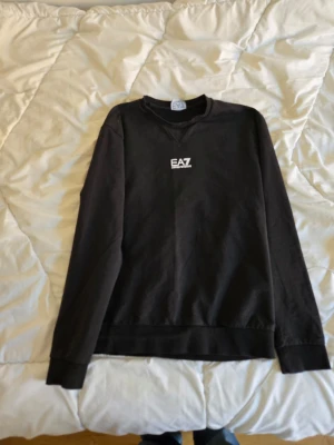 Svart sweatshirt från EA7 Emporio Armani - Svart sweatshirt med rund hals och vit EA7-logga på bröstet. Tröjan har långa ärmar och ribbade muddar vid ärmslut och nederkant. Perfekt för en stilren och avslappnad look. Se bilder för skick 