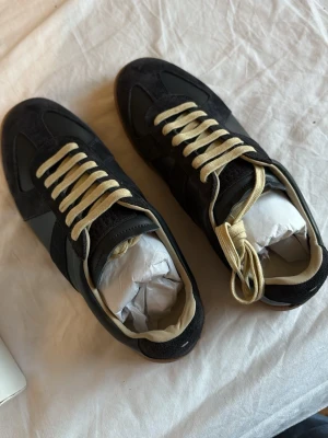 Maison Margiela Gats - Stilrena svarta sneakers från Maison Margiela med klassisk design, vita snören och kontrasterande gummisula. Ovandelen är i mocka och läder, vilket ger en lyxig känsla. Perfekta för dig som gillar minimalistisk och tidlös stil.