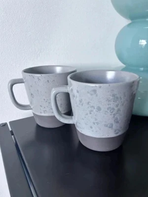Grå muggar från RAW Crafted Concept - Två stilrena grå muggar från RAW Crafted Concept med matt finish och subtilt prickigt mönster. Muggarna har breda handtag och en mörkare grå botten. Perfekta för kaffe eller te och passar snyggt i moderna hem.