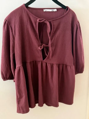 Vinröd blus med knytning - Vinröd blus från Lager 157 med knytning framtill. Blusen har en bekväm passform och är tillverkad i ett lätt, strukturerat tyg som ger en skön känsla. Perfekt för dig som gillar en avslappnad och trendig look.