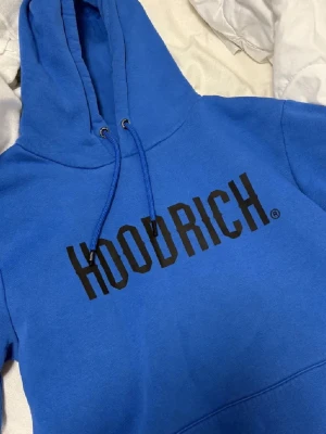 Blå HOODRICH hoodie med tryck - Snygg blå hoodie från HOODRICH med stor svart logga tryckt över bröstet. Klassisk modell med huva och dragsnören. Tillverkad i mjukt material som känns skönt mot huden. Perfekt för dig som gillar streetwear och vill sticka ut.