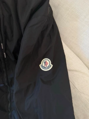 Svart Moncler vindjacka med huva - Snygg svart vindjacka från Moncler med huva och dragkedja. Jackan har Moncler-logga på ärmen och vita knappar på fickorna. Huvan har en cool detalj med röd, vit och blå rand. Perfekt för blåsiga dagar och streetstyle.