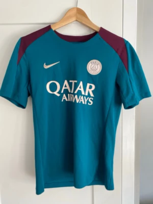 PSG Nike fotbollströja turkos - Snygg turkos PSG fotbollströja från Nike med vinröda axelpartier och vita tryck. Framsidan har Qatar Airways-logga och PSG-emblem, baksidan har Visit Rwanda-tryck. Perfekt för fotbollsträning eller att bära till vardags. Träningströja åt 2024 tror jag.