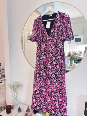 Blommig långklänning med v-ringning - Långklänning från H&M med v-ringning och korta puffärmar. Klänningen har ett färgstarkt blommönster i rosa, lila och vitt på svart botten. Tillverkad i lätt polyester, perfekt för sommarens alla tillfällen.