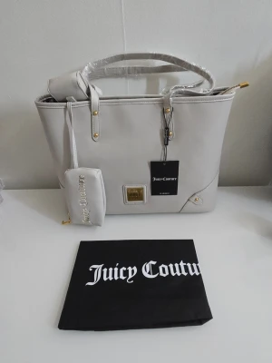 Ljusgrå handväska från Juicy Couture - Stilren ljusgrå handväska från Juicy Couture i slätt konstläder med guldfärgade detaljer. Väskan har matchande accessoarer, dragkedja och innerfoder med logotypmönster. Perfekt för dig som gillar trendiga och praktiska väskor.