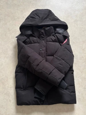 Svart dunjacka från Canada Goose - Säljer en svart dunjacka från Canada Goose med huva och klassisk logga på ärmen. Jackan har quiltad design, dragkedja och knappar framtill samt ribbade muddar vid ärmslut. Perfekt för kalla vinterdagar. Det står att den är storlek XS men den kan passa dig som har M/L